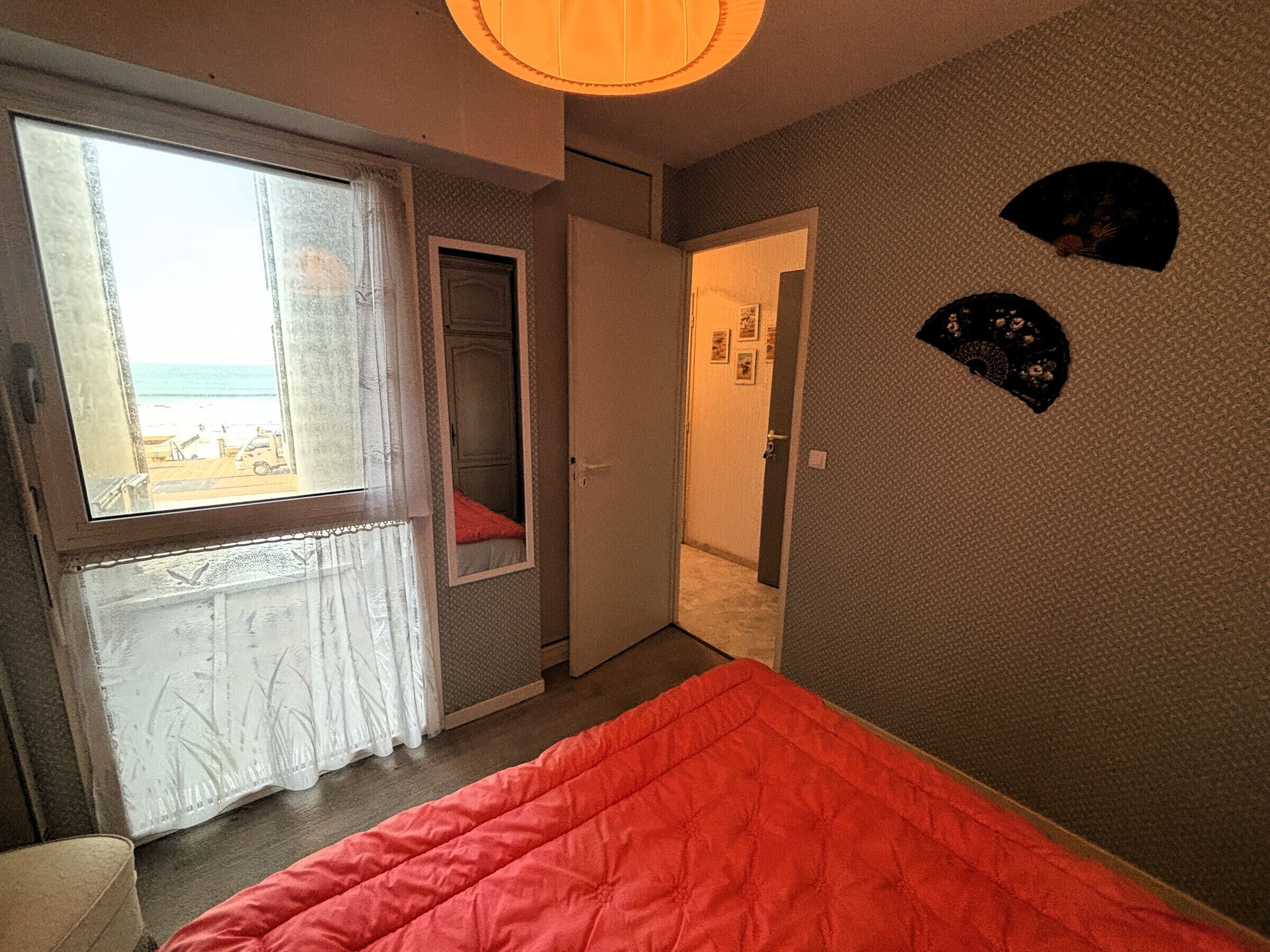 A bedroom with a large window, a bed with a red blanket, a door leading to a bathroom, and a wall with two decorative items.