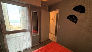 Un dormitorio con un ventanal grande, una cama con una colcha roja, una puerta que da al baño y una pared con dos adornos.