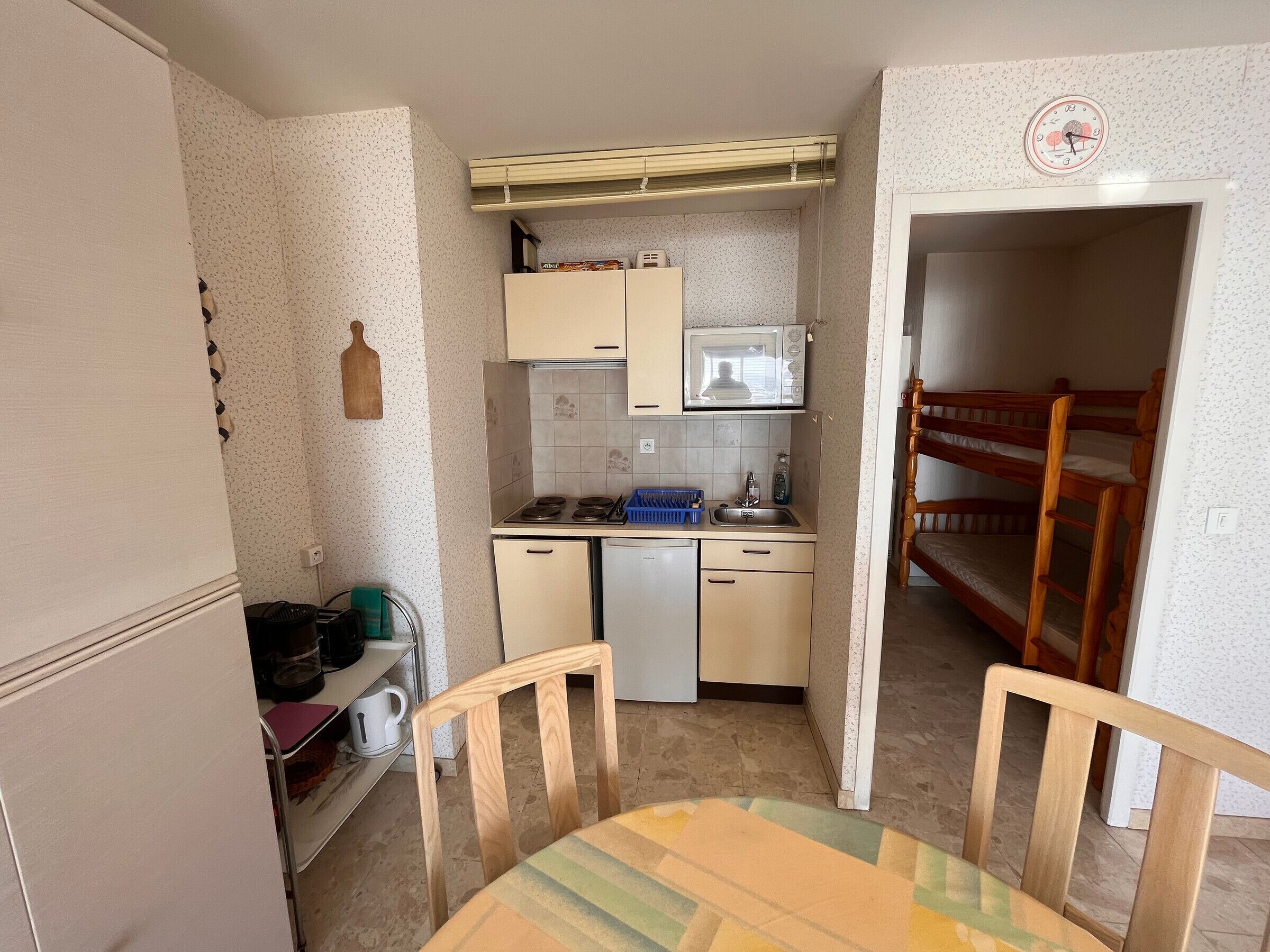 A compact kitchen with a microwave, sink, and a small refrigerator. There is a wooden chair and a table with a colorful tablecloth.