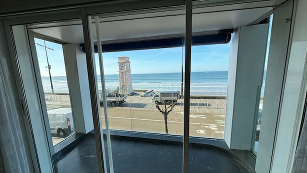 Vista desde una habitación con un ventanal grande que da a una playa, una torre del reloj y una carretera con vehículos.