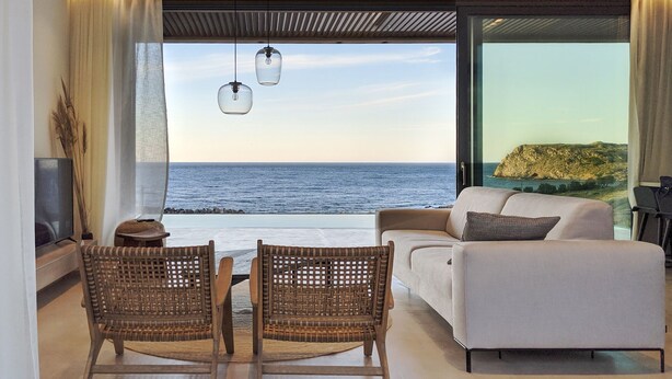 A modern living room with a sofa, two wicker chairs, a TV, and a view of the sea.