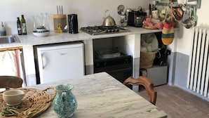 Een kleine keuken met een gasfornuis, een spoelbak en een kleine koelkast. Er staat een eettafel met een kom en een kan erop.