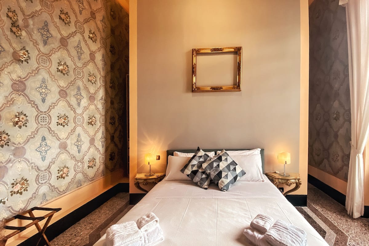 A hotel room with a large bed, two bedside lamps, patterned wallpaper, a mirror on the wall, and towels on the bed.