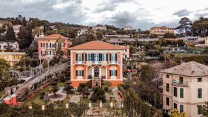 A large, two-story building with a red-tiled roof and green shutters, surrounded by lush gardens and a hillside residential area.