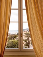 Una finestra con tende gialle, vista su una zona costiera con edifici e palme.