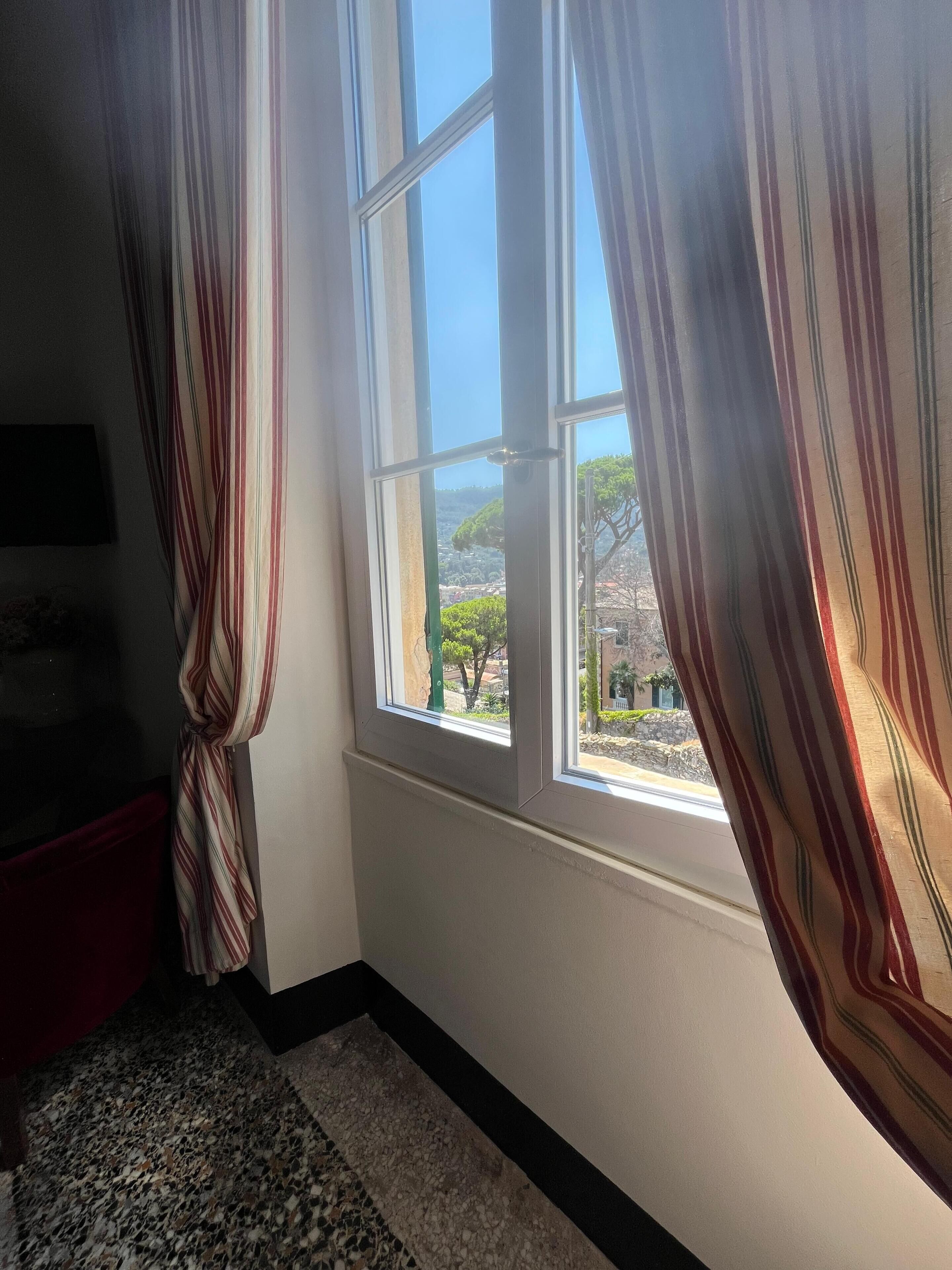 A room with a large window, striped curtains, a granite floor, and a view of a landscape with trees and buildings.