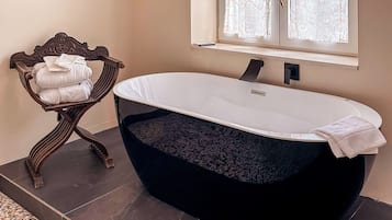 Un baño con una bañera independiente, una ventana con cortinas, un cuadro enmarcado en la pared y una silla decorativa con toallas.