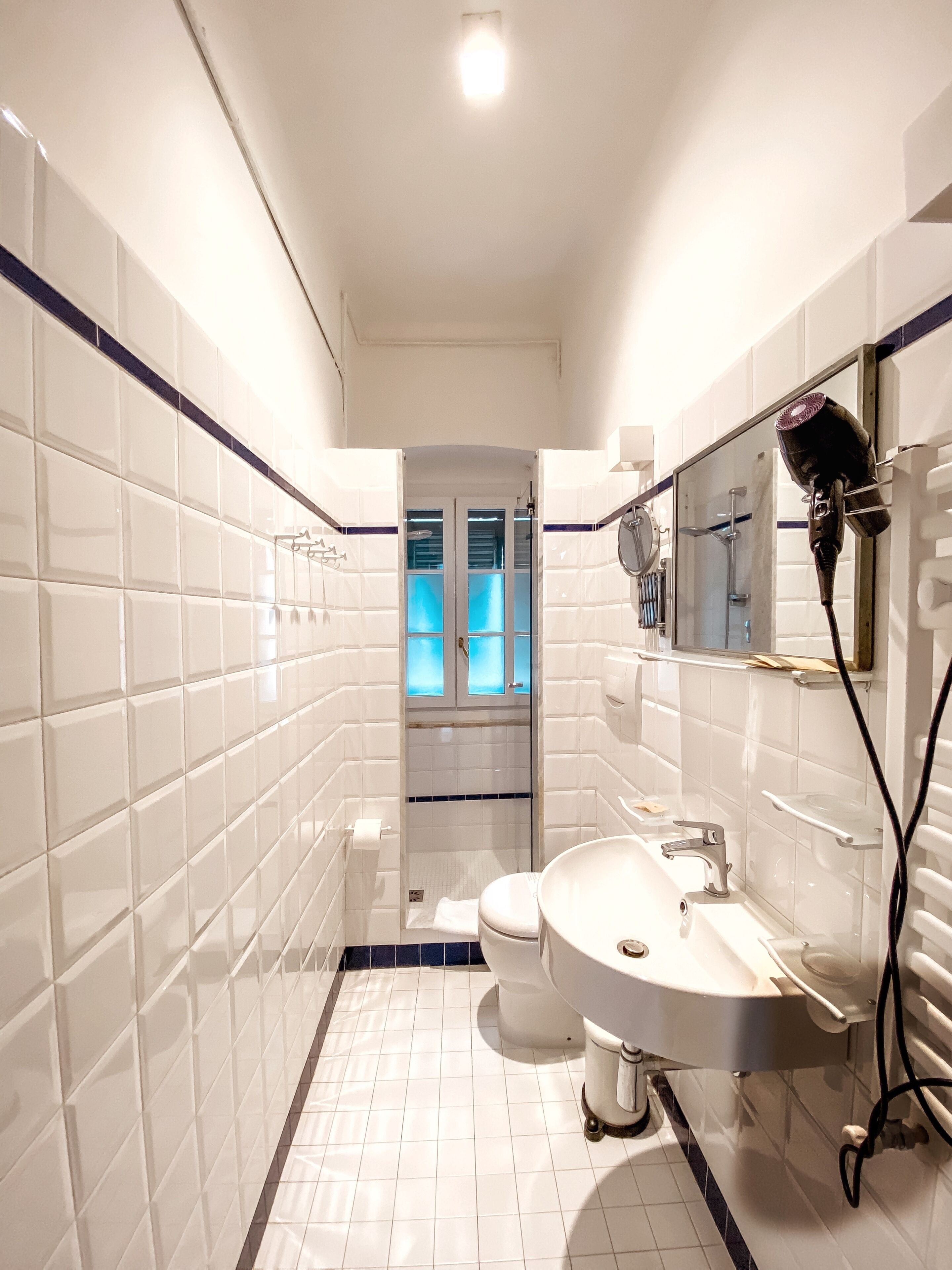 Une salle de bain avec des murs recouverts de carreaux blancs, un lavabo blanc, un miroir et un sèche-cheveux.