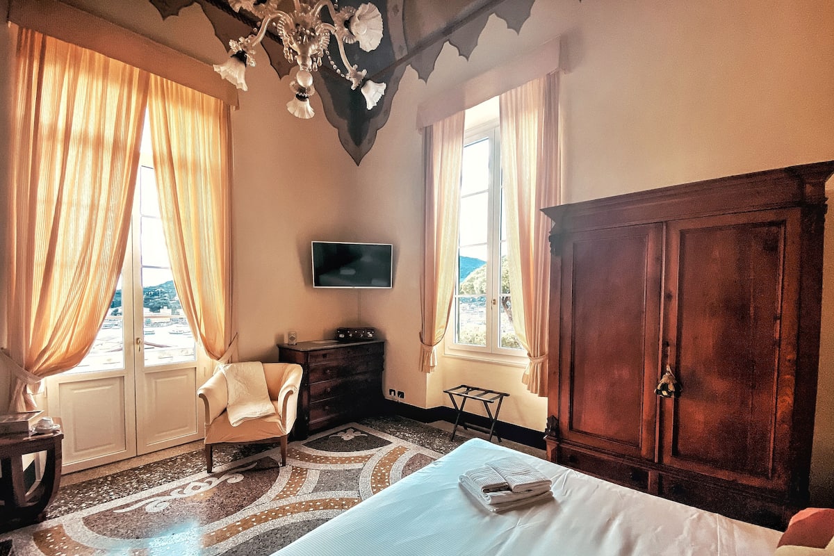 A bedroom with a large bed, a wooden wardrobe, a television on the wall, and a view of the outdoors through the window.