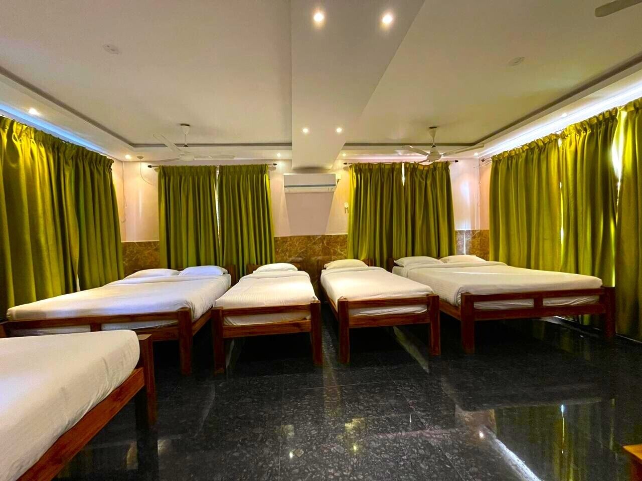 Habitación de hotel con cuatro camas individuales, cortinas verdes, piso de mármol y ventilador de techo.