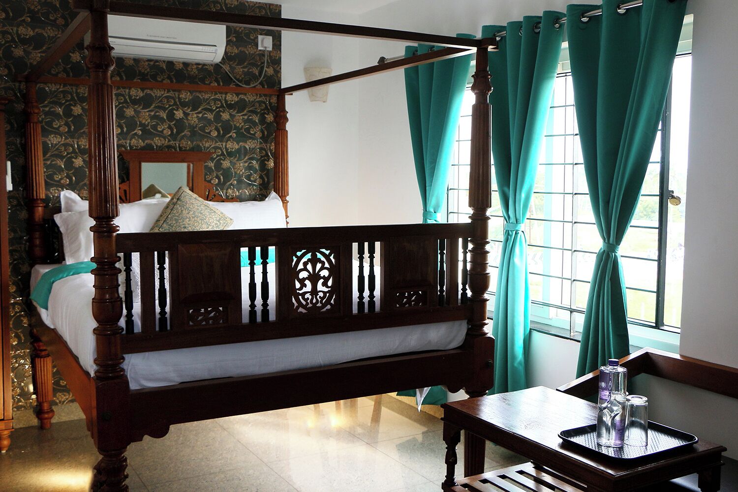 A four-poster bed with a wooden frame and a canopy, a small wooden table with glasses, and a window with curtains.