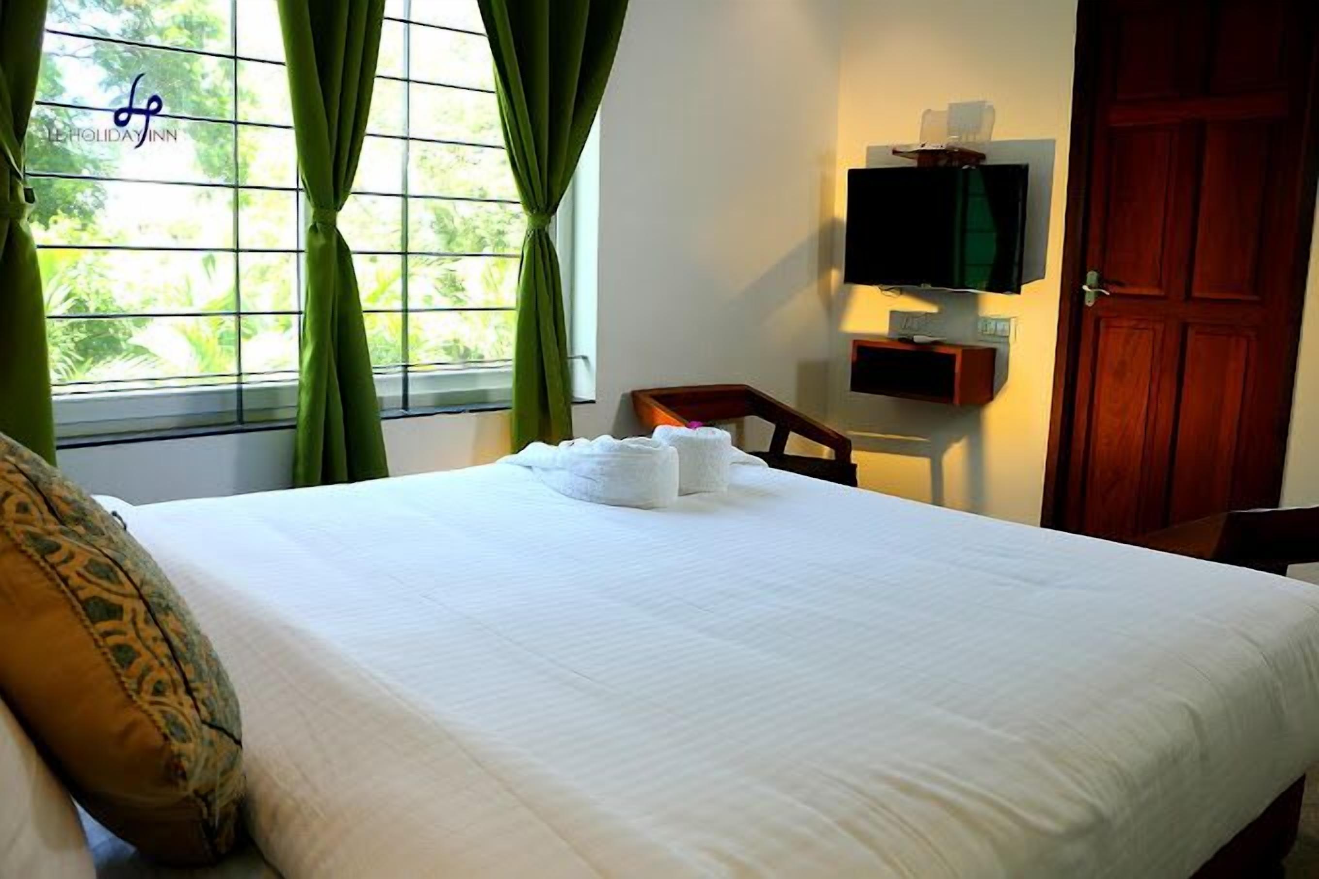 Habitación de hotel con cama, televisor montado en la pared, puerta de madera y ventana con cortinas.