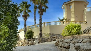 A paved pathway leading to a building with a beige facade, palm trees, and a stone wall. - Luxury villa with sea view, with Swimming pool (Campello)