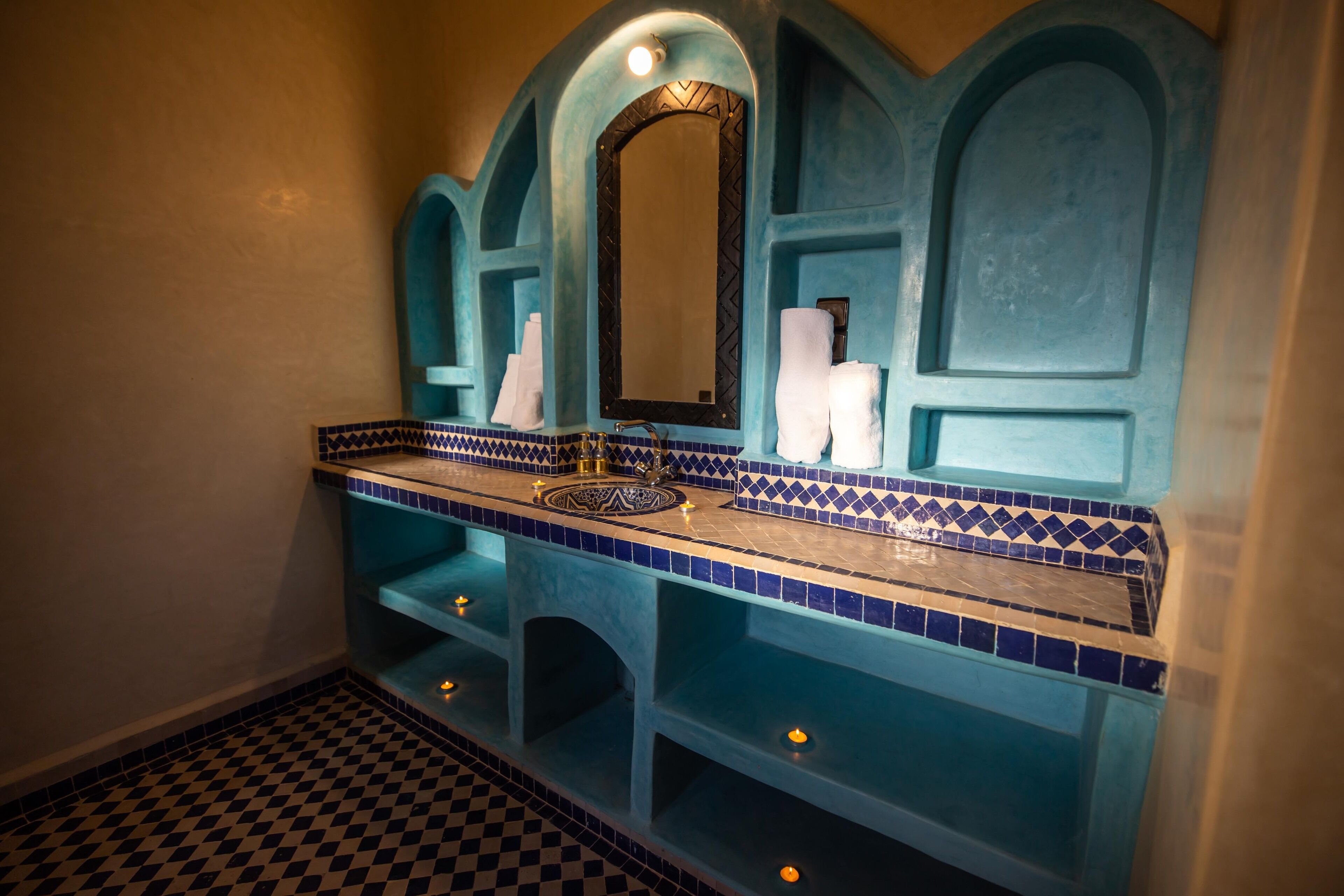 Un baño con piso a cuadros, un lavamanos con salpicadero de azulejos azules y blancos, y un espejo con marco negro.