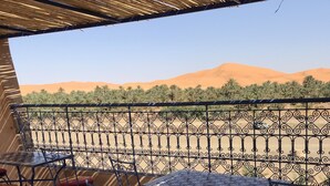 Une terrasse extérieure avec des tables et des chaises en métal, un plafond décoré et une vue sur des palmiers et des dunes de sable.