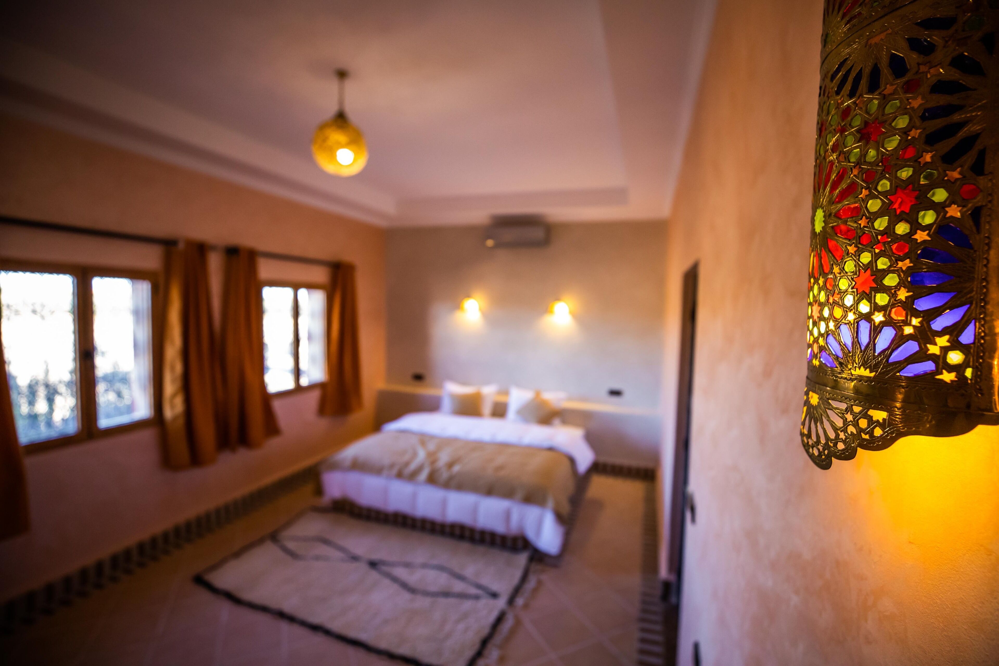 Une chambre à coucher avec un lit, une table de chevet, une lampe fixée au mur et une fenêtre avec des rideaux.
