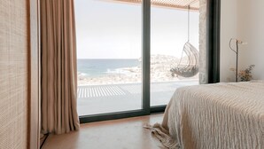 Un dormitorio con un ventanal grande con vista a la playa, un sillón colgante y una cama con un edredón texturizado.