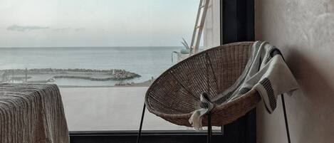 A room with a large window overlooking a beach, a wicker chair, and a ladder leaning against the wall.