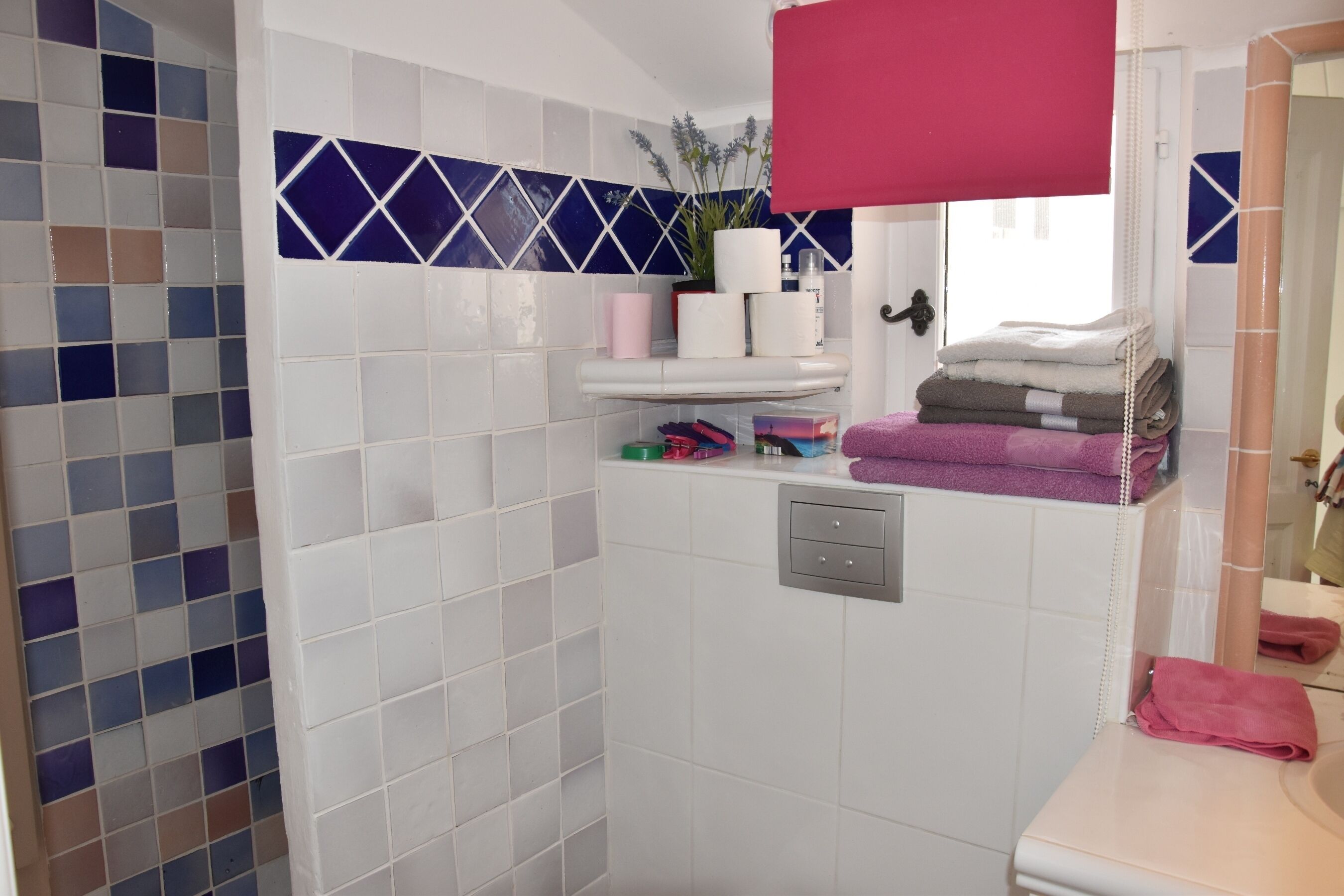 Casa de banho com azulejos brancos, um painel decorativo de mosaico azul e branco, uma toalha cor-de-rosa e um espelho.