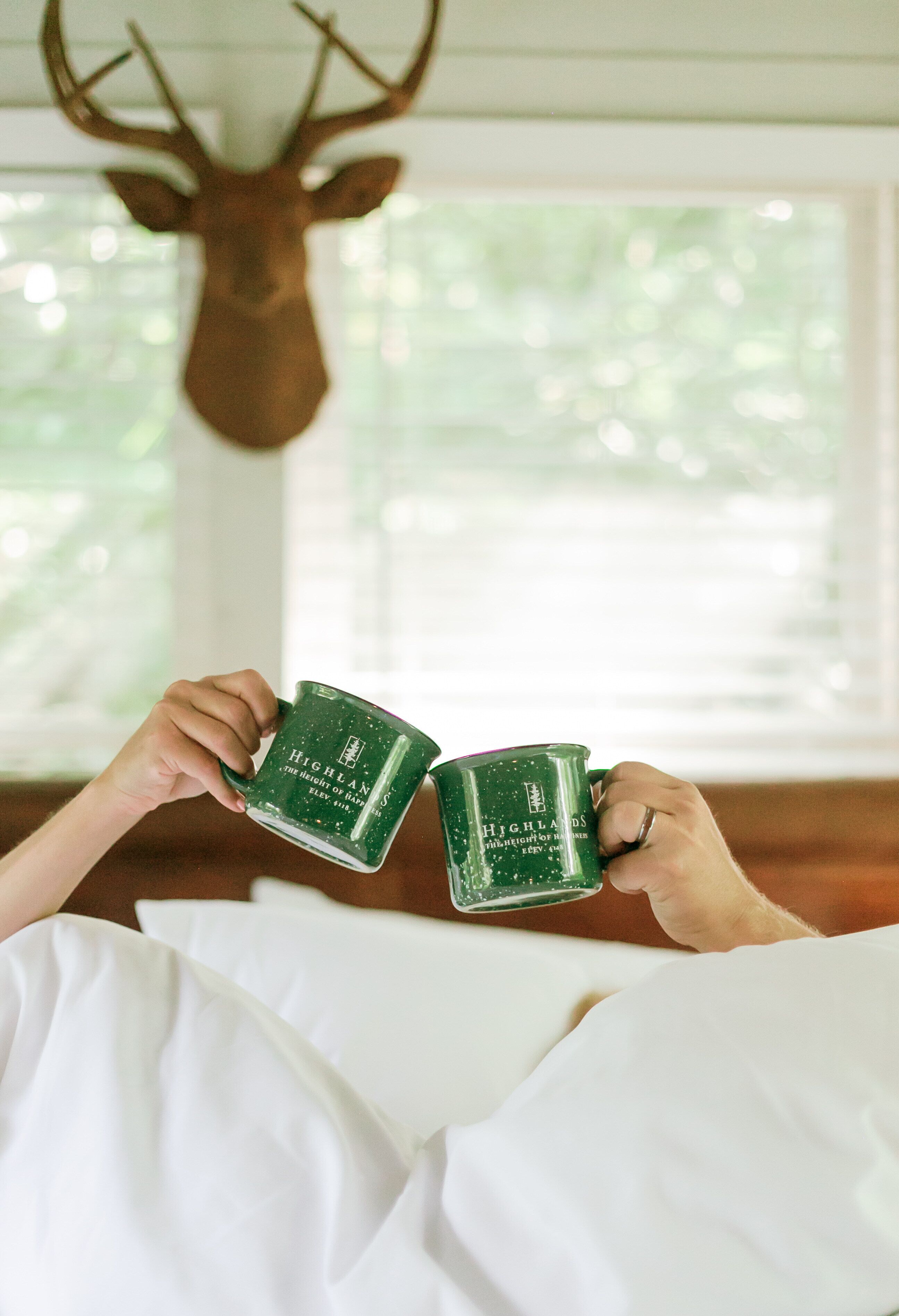 A person holding two green mugs with "Highlands" text, sitting in a room with a deer head mounted on the wall and a window with blinds.