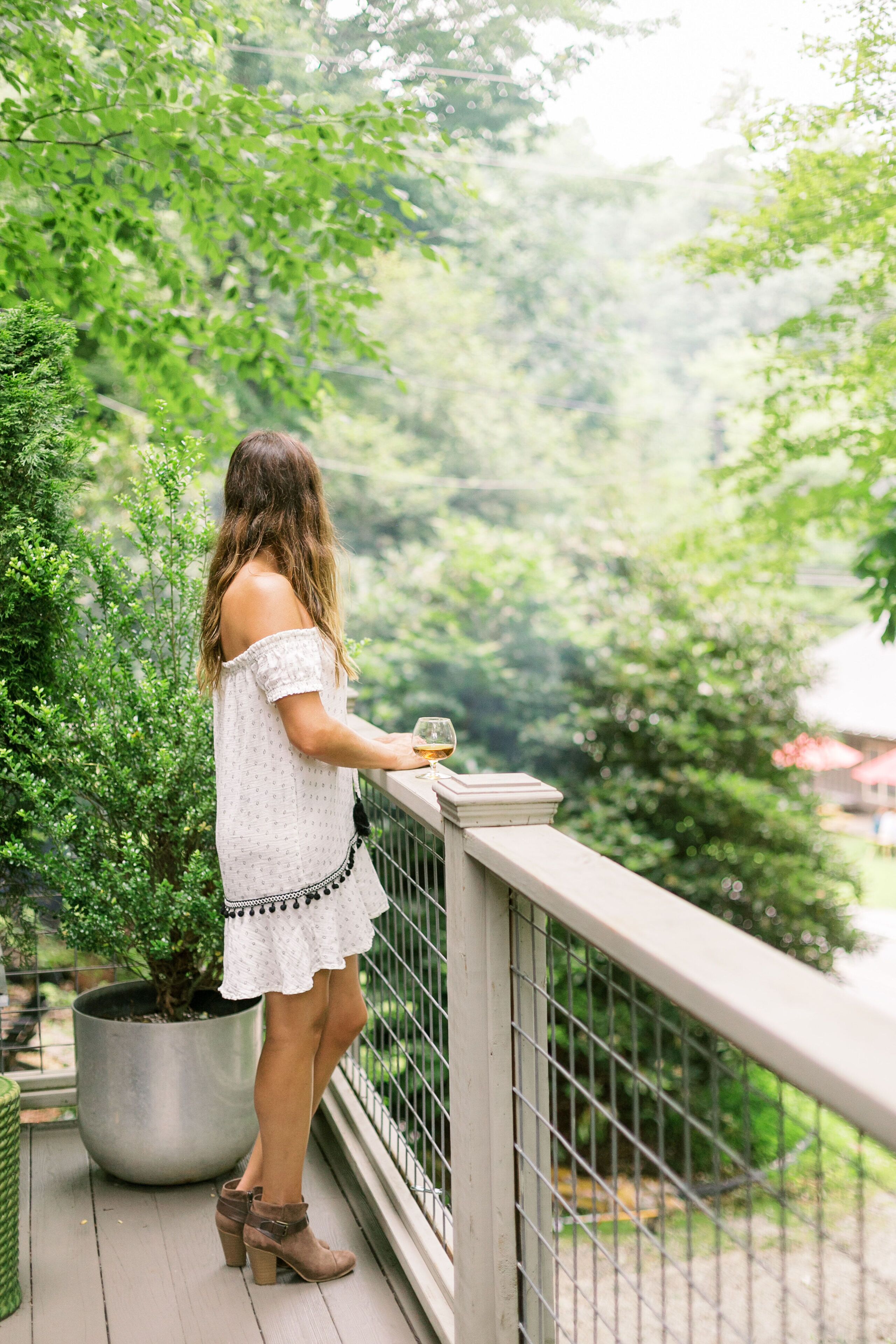 Une personne se tient sur un balcon doté d’une balustrade, elle tient un verre à la main et regarde un jardin avec des arbres et une plante en pot.