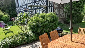 Un tavolo e sedie in legno sotto un ombrellone, un barbecue e un giardino con siepe e una sedia.