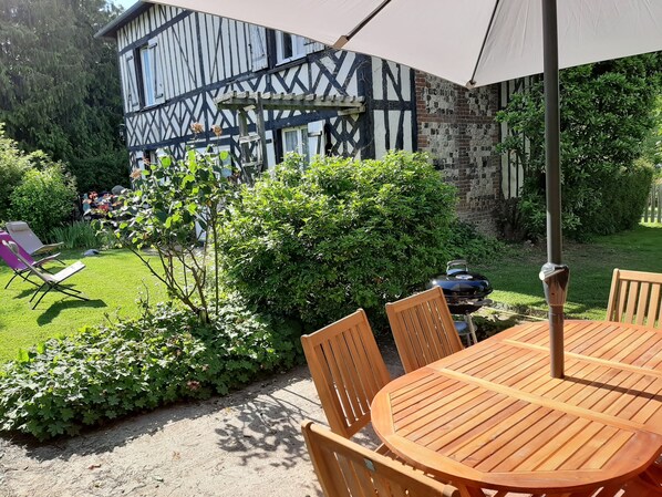 A wooden table and chairs under an umbrella, a barbecue grill, and a garden with a hedge and a chair.