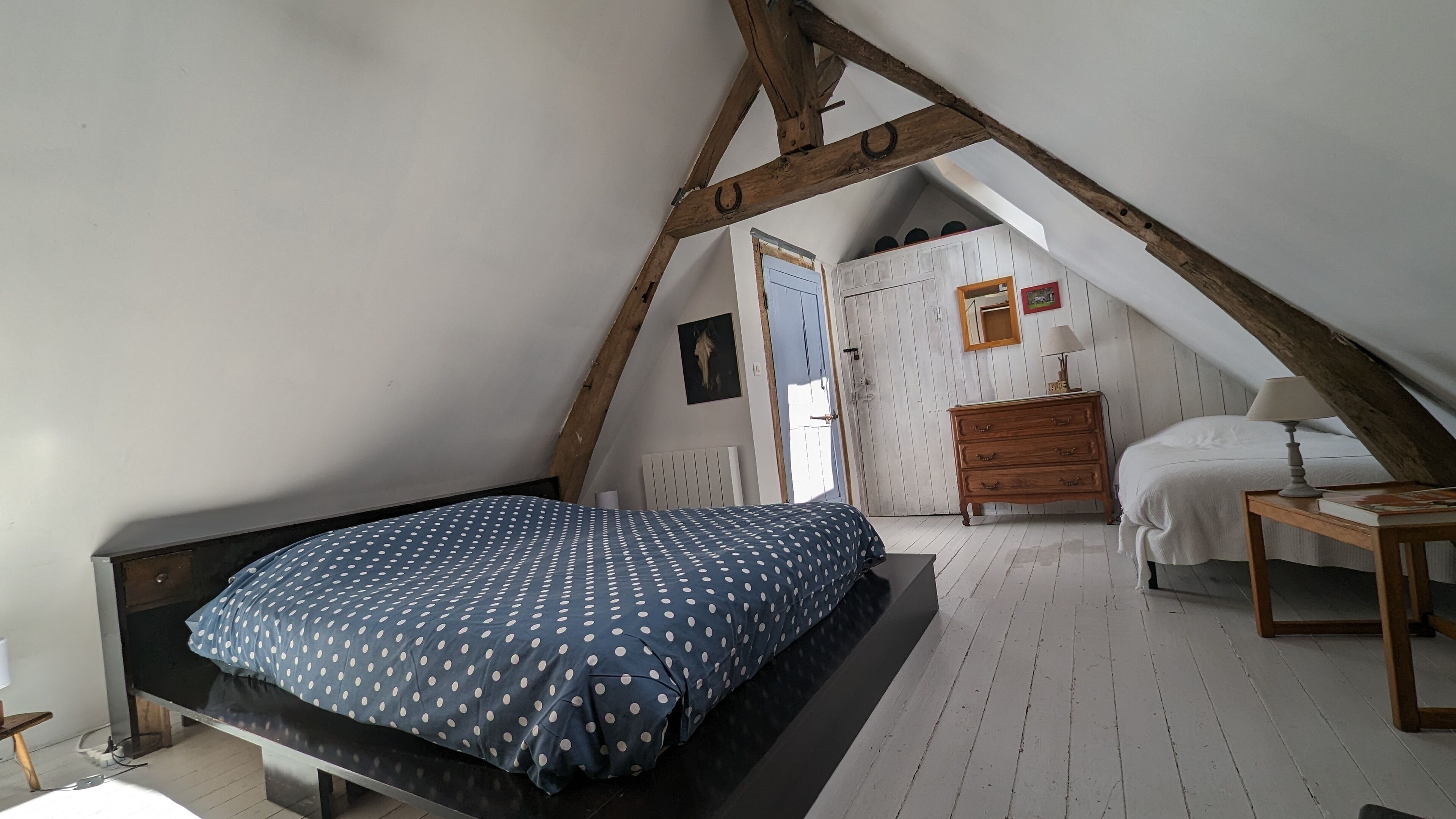 Un dormitorio con techo inclinado, una cama con colcha de lunares, un aparador de madera, una mesita de noche con lámpara y una ventana con vistas al exterior.