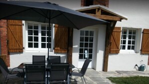 An outdoor patio with a table and chairs under a large umbrella, a stone floor, and a white building with wooden shutters.