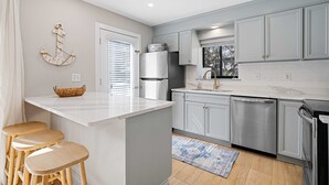 A modern kitchen with grey cabinets, a white countertop, and a stainless steel refrigerator. There are three wooden stools at the island, and a window with blinds. - Newly Renovated Luxury Villa- 5 minute walk to Beach! (Hilton Head Island)