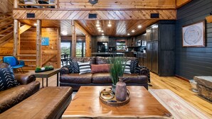 Un vaste espace de vie doté d’un haut plafond, de poutres apparentes, d’une cheminée, de canapés en cuir, d’une table basse en bois et offrant une vue sur l’extérieur.