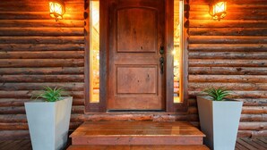 L’entrée d’un chalet en bois, avec une porte à deux battants, deux lanternes fixées au mur et deux grands jardinières contenant des plantes vertes.