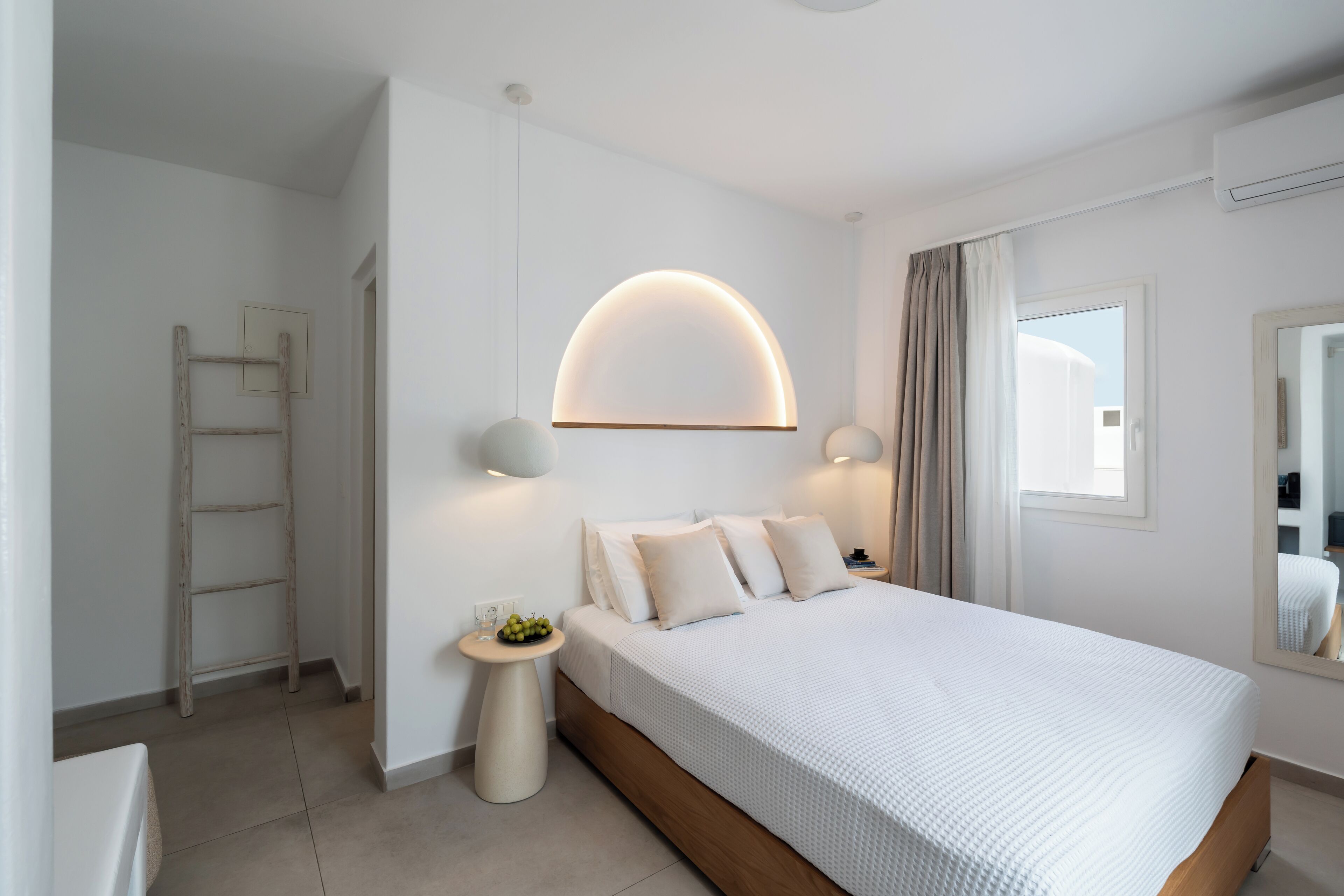 A modern bedroom with a large bed, bedside lamps, a small round table with a fruit bowl, and a ladder against the wall.