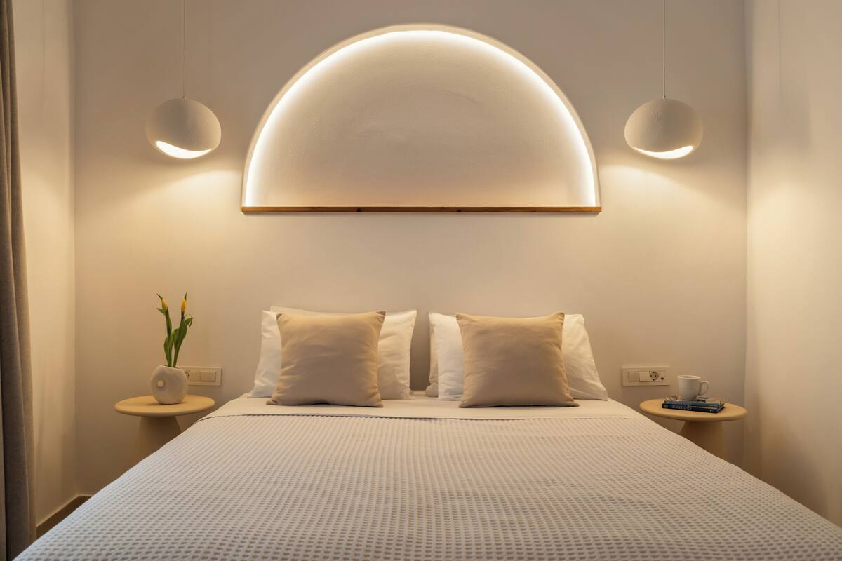 A neatly made bed with a large, semi-circular light fixture above it, two bedside tables with a vase of flowers and a cup, and a wall outlet.