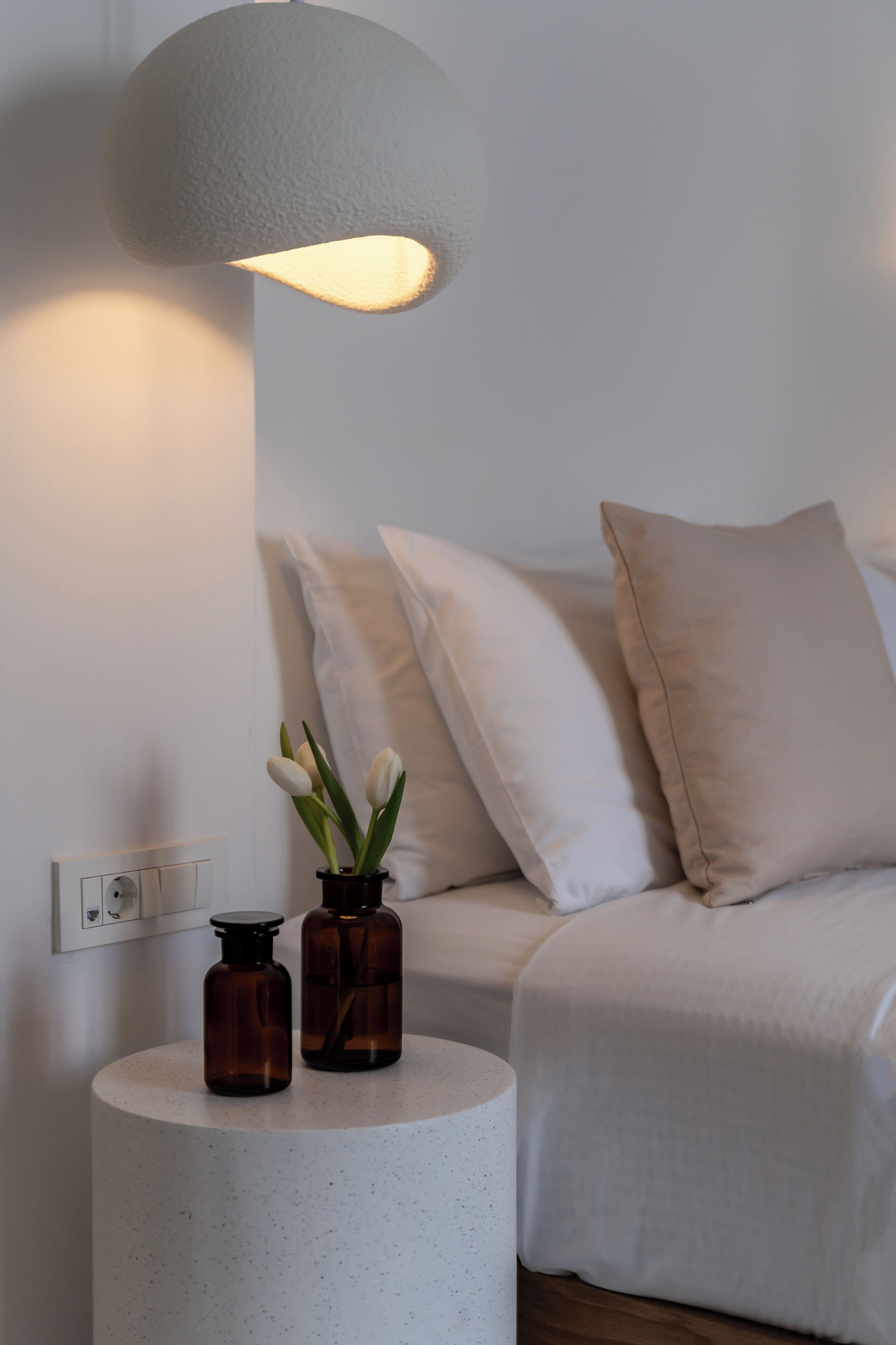 Une chambre à coucher avec une table d’appoint ronde, deux bouteilles marron et un vase avec des fleurs blanches.