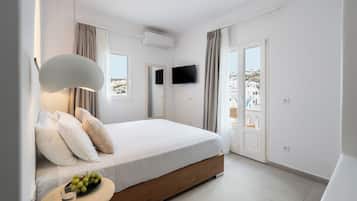 A bedroom with a bed, a bedside table with a bowl of fruit, a TV, and a view of buildings through the window.