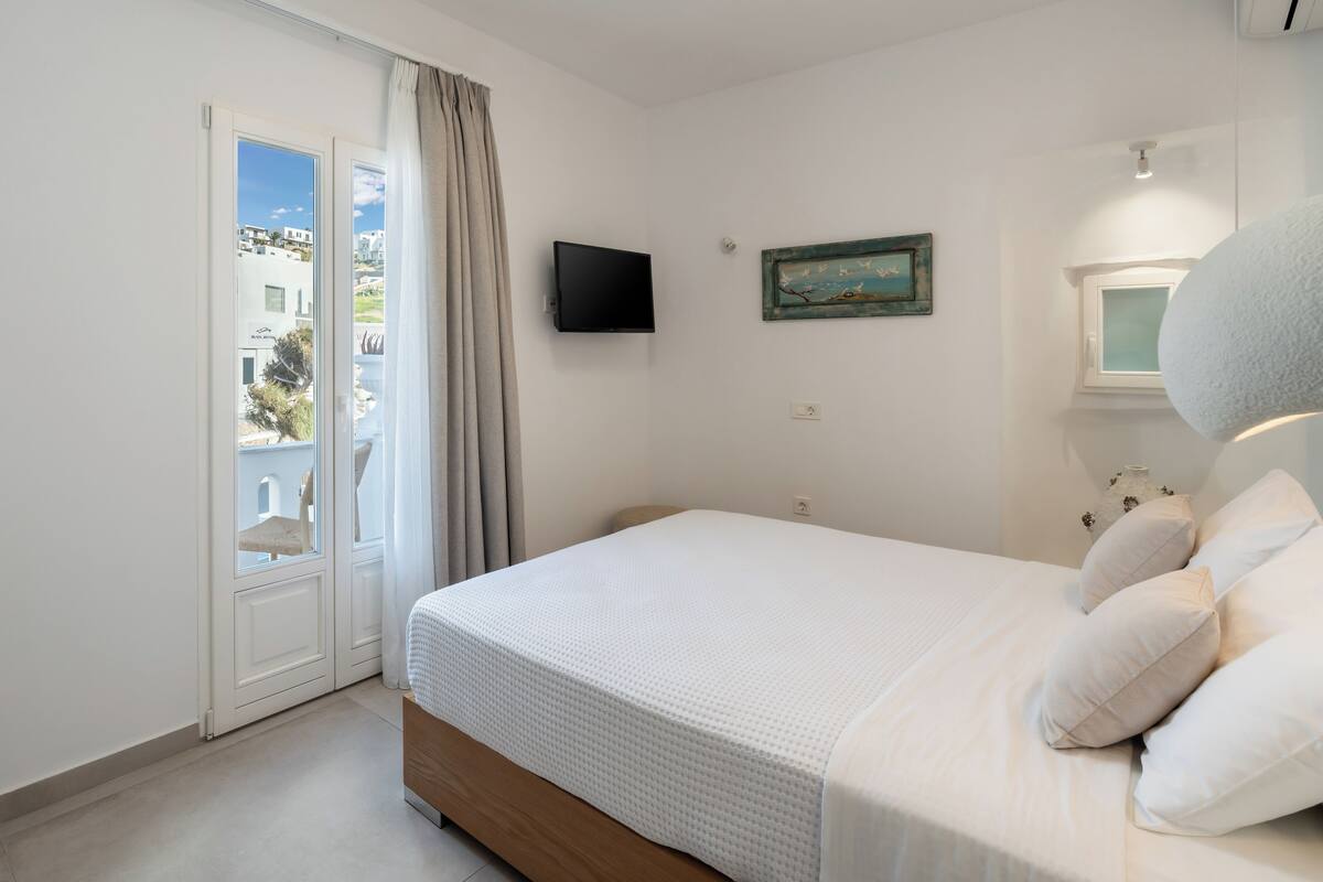 A bedroom with a bed, a television, a window, and a view of buildings outside.