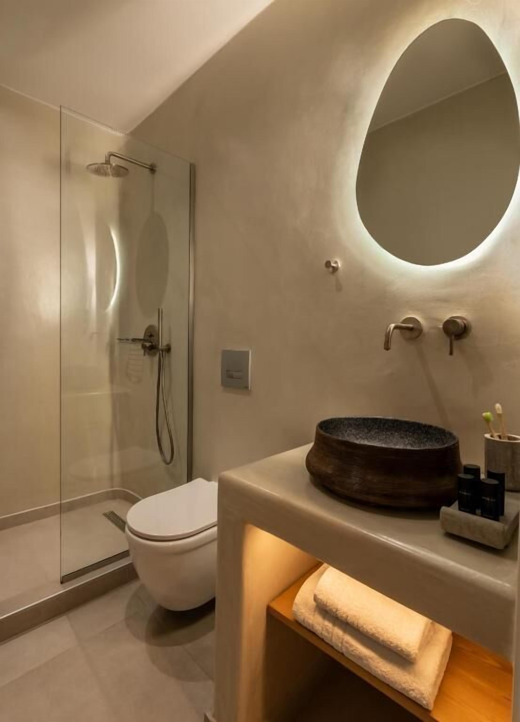 A modern bathroom with a glass shower enclosure, a white toilet, and a round mirror above a sink with a wooden basin.