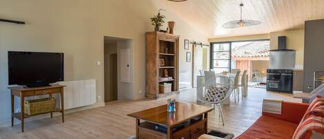 A spacious living area with a wooden ceiling, a television, a wooden coffee table, a red sofa, and a kitchen in the background.