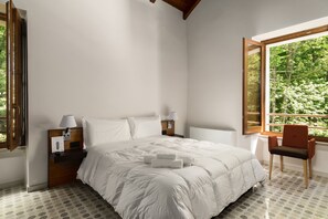 A bedroom with a large bed, two bedside tables with lamps, a chair, and a window with a view of greenery. - Elda Hotel (Monte Sant'Angelo)