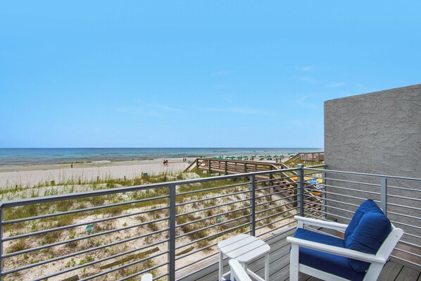 A balcony with a view of the beach, a chair with a cushion, and a railing.