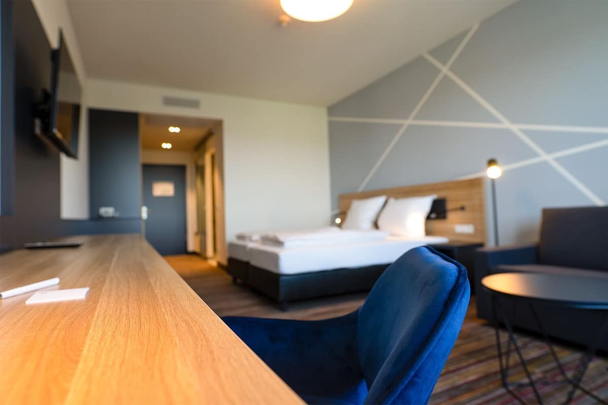Una habitación de hotel con un escritorio de madera, una cama con ropa blanca, una silla azul y una pequeña mesa redonda.