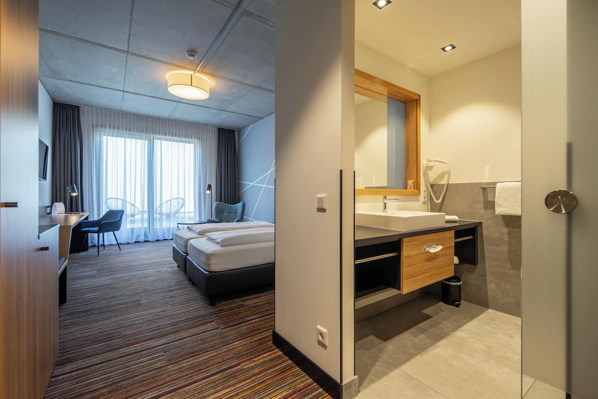 Habitación de hotel con cama, escritorio, silla, baño con lavamanos y espejo, y ventana con cortinas.