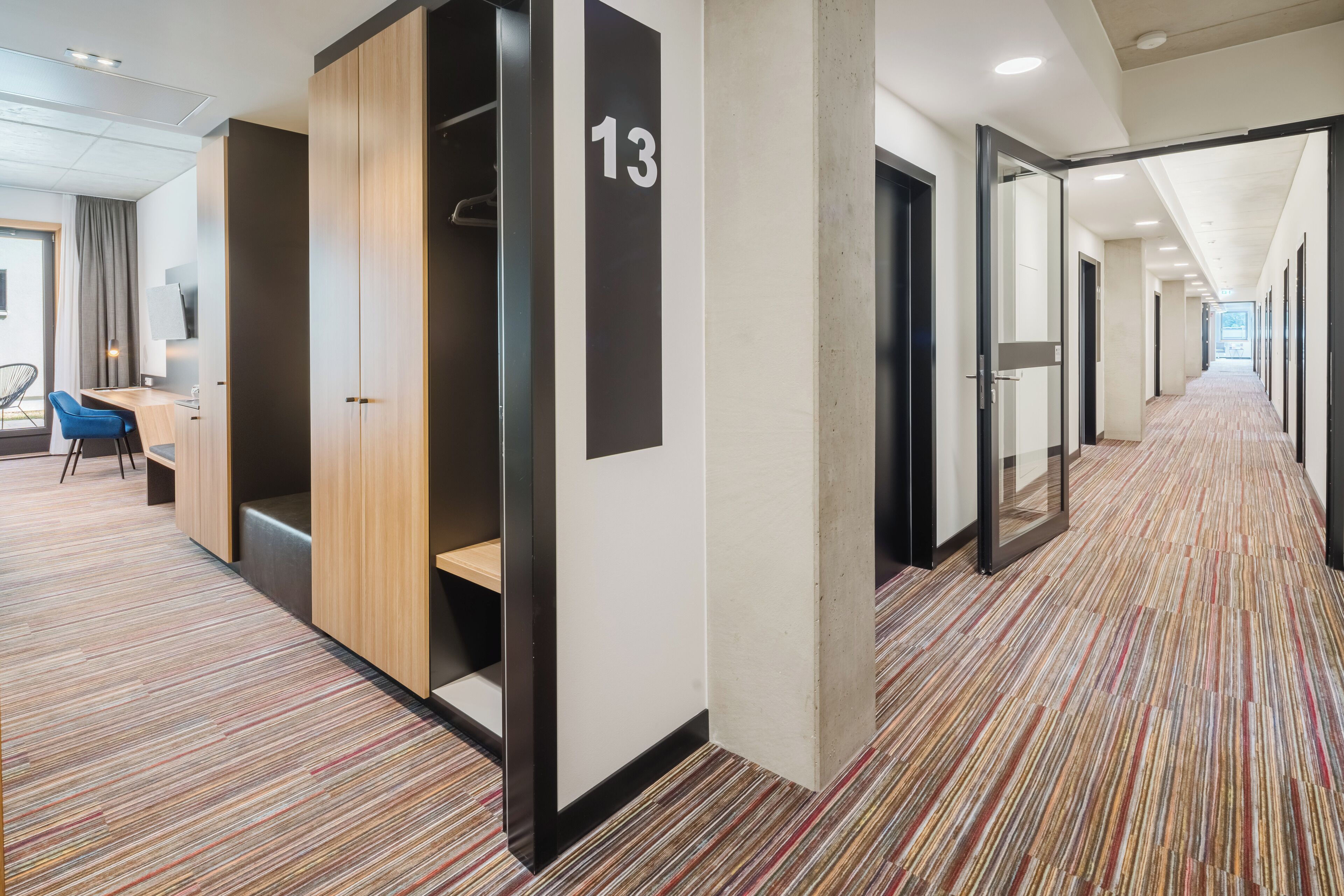 Un pasillo moderno de hotel con alfombra a rayas, un escritorio con sillas y un cartel con el número 13 en la pared.