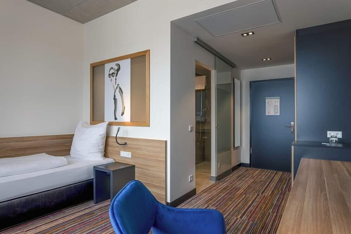 Habitación de hotel con cama, escritorio, silla, un cuadro en la pared y una puerta que da al baño.
