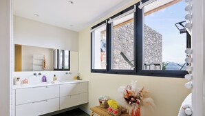 A bathroom with a large mirror, white cabinets, a wicker stool, and a view of a stone building through a window.