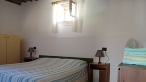 A bedroom with a bed, two bedside tables, a wardrobe, a window with curtains, and a fan on a wooden cabinet. - Two-room apartment with garden and private parking space (Portoferraio)