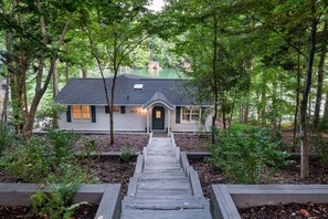 Une maison dotée d  ’un porche à l’avant, entourée d’arbres et accessible par un chemin en bois menant à l’entrée.
