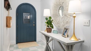Un corridoio con una porta blu, una parete bianca con uno specchio decorativo, un tavolo con una lampada e una pianta.