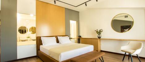 A modern hotel room with a wooden bed, a bedside table, a chair, a large mirror, and a bathroom with a sink and mirror.